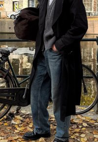 Svart lång kappa över en grå cardigan och vit skjorta, kombinerat med blå jeans och svarta skor. En brun väska hänger över ena axeln.