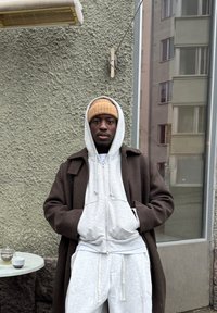 Personne portant un sweat à capuche gris, un pantalon de survêtement gris et un manteau marron, debout près d'un mur vert clair, avec un bonnet beige et des tasses sur une table à proximité.