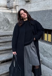 Cappotto lungo nero sopra un maglione a maglia nero, abbinato a pantaloni corti a righe. Porta una borsa a spalla nera e indossa stivali neri fino al ginocchio.