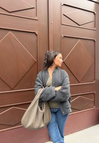 Grå polstret jakke over en grønn topp, blå jeans, holder en stor flettet tanfarget veske, står foran en brun bakgrunn med diamantmønster.