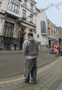 Κάζουαλ γκρι φούτερ και ξεθωριασμένα τζιν φορεμένα από ένα άτομο που είναι στραμμένο προς έναν δρόμο με ιστορικά κτίρια και φράκτες κατασκευών.