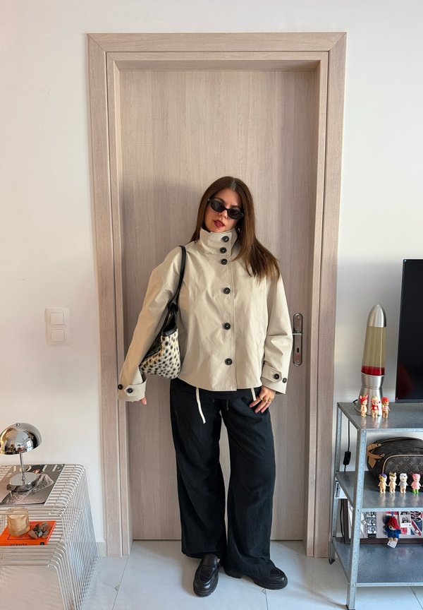 Una mujer con gafas de sol posa junto a una puerta de madera. Lleva una chaqueta color crema con botones, pantalones negros anchos y un bolso estampado al hombro.