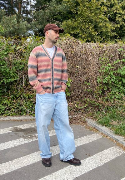 Hombre con cárdigan de rayas, vaqueros anchos azul claro, gorra marrón y zapatos oscuros de pie en un paso de peatones cerca de arbustos verdes.