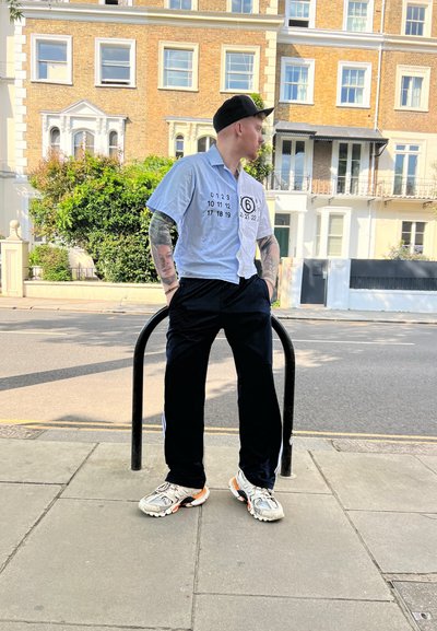 Camisa blanca de manga corta con números y logotipo en negro impresos, combinada con pantalones negros y zapatillas de correr plateadas con detalles en naranja.