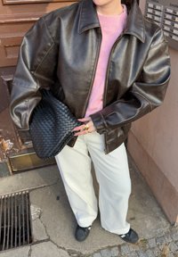 Brown leather zip-up jacket with a structured design, teamed with a pink sweater, white wide-leg trousers, and a black woven clutch.