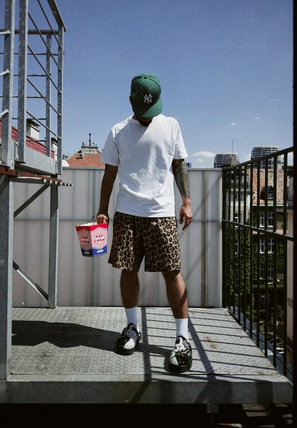 Camiseta branca de manga curta, calções de estampado de leopardo, sapatos preto e branco, segurando um balde de pipoca riscado em vermelho e azul, em pé numa varanda.