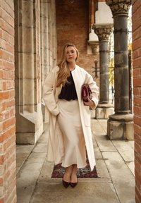 Bež dolg plašč čez črno majico, kombinirano s kremasto krilo in vinsko rdečimi čevlji z visoko peto. Obleka vključuje majhno rdečo torbico z gladkimi teksturami.