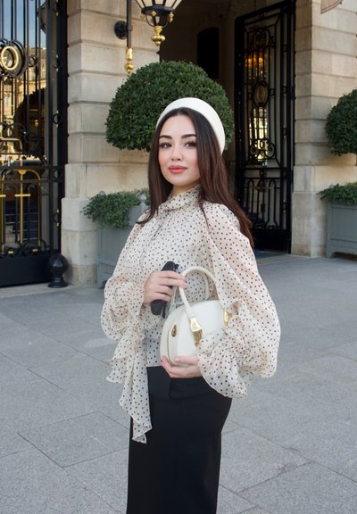 Joven mujer con blusa transparente de lunares y falda negra sostiene un bolso blanco, parada frente a un edificio elegante con plantas decorativas.