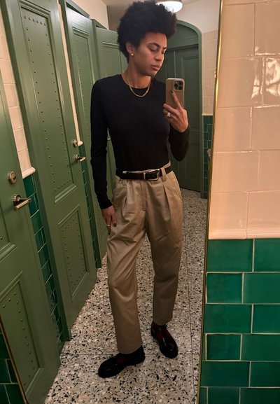 Camiseta negra de manga larga, pantalones de tiro alto beige, cinturón negro con hebilla de plata y zapatos oscuros con detalles rojos, de pie en un espacio con espejos.