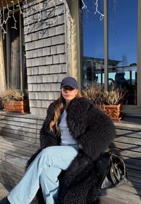 Femme portant un manteau en fourrure noire et une casquette bleu marine, assise sur un banc en bois devant un bâtiment avec des plantes en pot et de grandes fenêtres en verre.