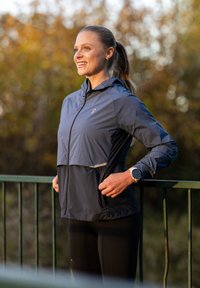 Naine spordijakiga ja retuusidega seisab õues piirde ääres, naeratab ja vaatab kõrvale kuldse tunni ajal.