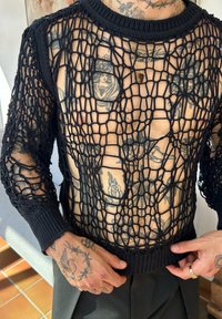 Svart stickad mesh-topp med ett stort diamantmönster, rund halsringning och ribbade ärmslut. Exponerar hud och tatueringar under.