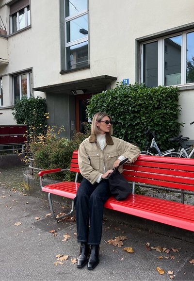 Chaqueta de pana beige con cuello marrón, suéter blanco, jeans oscuros y botas negras, sentado en un banco de color rojo brillante con vegetación cerca.