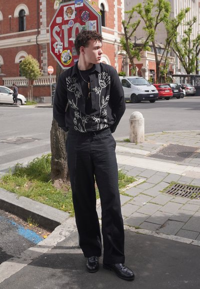 Camisa negra de botones con bordado floral en blanco, combinada con pantalones negros de pierna ancha y zapatos negros brillantes, de pie cerca de una señal de alto.