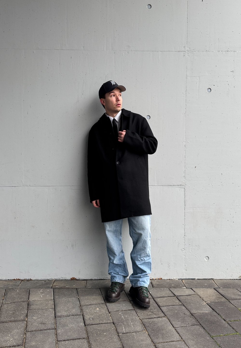 Manteau en laine noir avec un seul bouton, superposé à une chemise blanche et une cravate noire, associé à un jean bleu clair et des bottes noires. Modèle masculin jeune adulte.