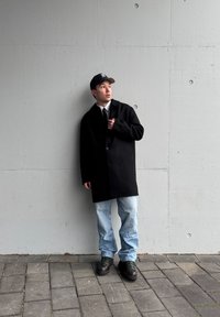 Manteau en laine noir avec un seul bouton, superposé à une chemise blanche et une cravate noire, associé à un jean bleu clair et des bottes noires. Modèle masculin jeune adulte.