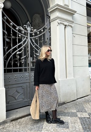 Mujer con suéter negro, falda de leopardo y gafas de sol se encuentra cerca de una reja de hierro ornamentada, sosteniendo una bolsa beige en una acera de adoquines.