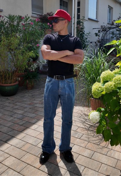 Gorra roja, camiseta negra, jeans azules y zapatos negros. De pie con los brazos cruzados en un jardín con plantas verdes y flores.