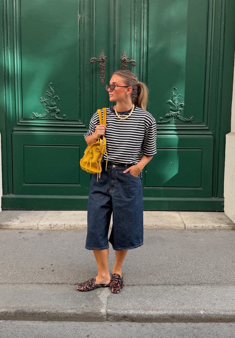 Femme portant un t-shirt blanc et noir rayé et un short en jean large, se tenant devant de grandes portes doubles vertes ornées avec un sac à main jaune.