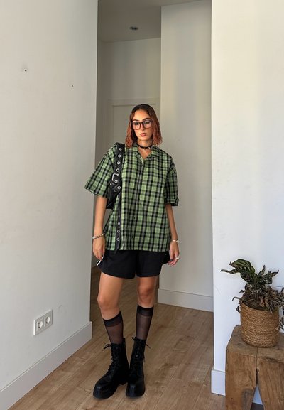Camisa de cuadros verde de gran tamaño con cuello, combinada con pantalones cortos negros. Botas negras de cordones y medias altas transparentes. Bolso cruzado negro.