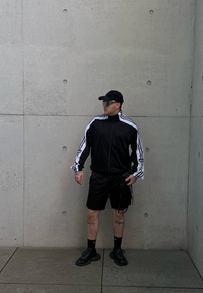 Hombre con chándal Adidas negro y blanco, gorra negra, gafas de sol y zapatillas, está apoyado en una pared de hormigón gris sosteniendo una cámara.
