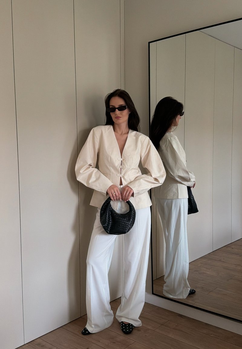 Mujer con blazer crema grande y pantalones blancos, sosteniendo un bolso tejido negro, de pie cerca de un espejo de cuerpo entero sobre suelo de madera.