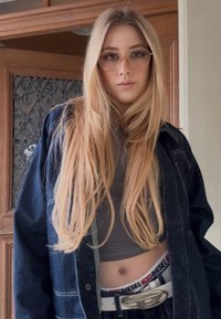 Jeune femme aux longs cheveux blonds, portant des lunettes, une veste sombre, un haut court, et une ceinture Diesel visible au-dessus de la ceinture du jean.