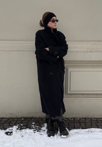 Manteau en laine noir surdimensionné avec un col montant et deux boutons à l'avant. Porté avec un pantalon gris et des bottes noires à lacets. Fond de neige et pavés.