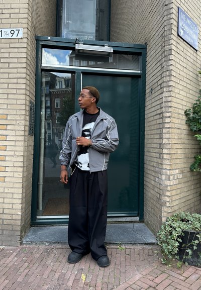 Rompevientos gris con cremallera, corte relajado, detalles en blanco; camiseta gráfica negra debajo; pantalones negros holgados; entorno al aire libre.