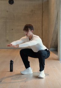 Inimene, kes kannab valget tihedat pika varrukaga pusa ja musti pükse, kükitab puidust põrandal, tema kõrval on must joogipudel.