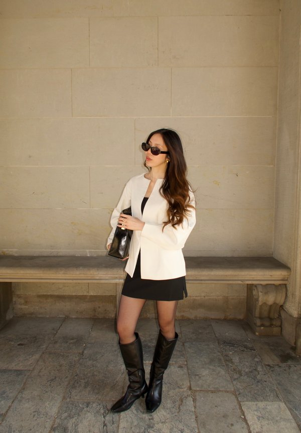 Witte blazer, zwarte mini-jurk, zwarte kniehoge laarzen. Accessoires zijn een zwarte clutch en grote zonnebril. Textuur achtergrond.
