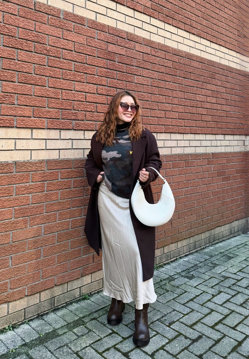 Femme portant des lunettes de soleil, un manteau sombre, un pull camouflage, une jupe beige, des bottes marron, tenant un sac à main blanc en forme de croissant, debout sur un trottoir pavé près d'un mur en briques.