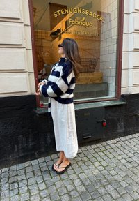 Marineblauer und weißer kariert gestrickter Pullover, weißes fließendes Kleid mit Spitze am Saum, schwarze Flip-Flops; Person steht am Fenster einer Bäckerei.