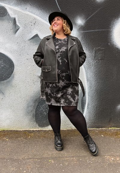 Chaqueta de cuero negra sobre un vestido gris con efecto tie-dye, medias negras y botas negras con cordones. El fondo presenta un mural en tonos gris y negro.