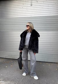 Casaco de pêlo sintético preto com um colarinho largo, suéter cinza, jeans cinza claro e tênis brancos; segurando uma bolsa tote preta.