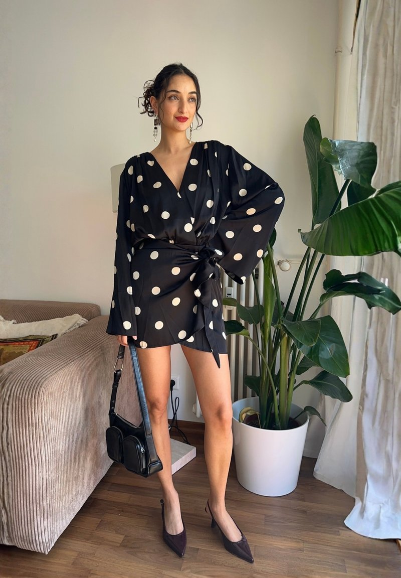 Donna con capelli ricci vestita con un abito a portafoglio nero a pois e tacchi, tiene una borsa nera in mano, in piedi accanto a una grande pianta in vaso in soggiorno.