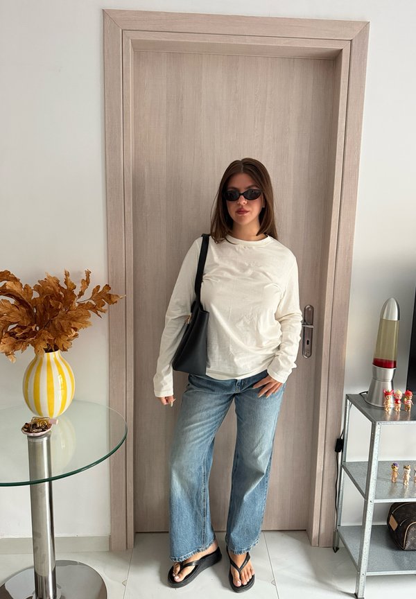 Mujer con gafas de sol, camisa blanca de manga larga, vaqueros azules y sandalias negras está de pie sosteniendo una bolsa negra delante de una puerta de madera.