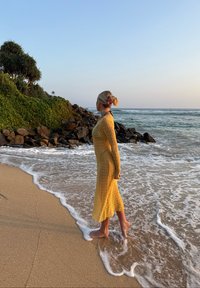 Femme en robe jaune debout pieds nus sur une plage de sable avec les vagues qui arrivent, faisant face à un littoral rocheux couvert de végétation verte sous un ciel dégagé.