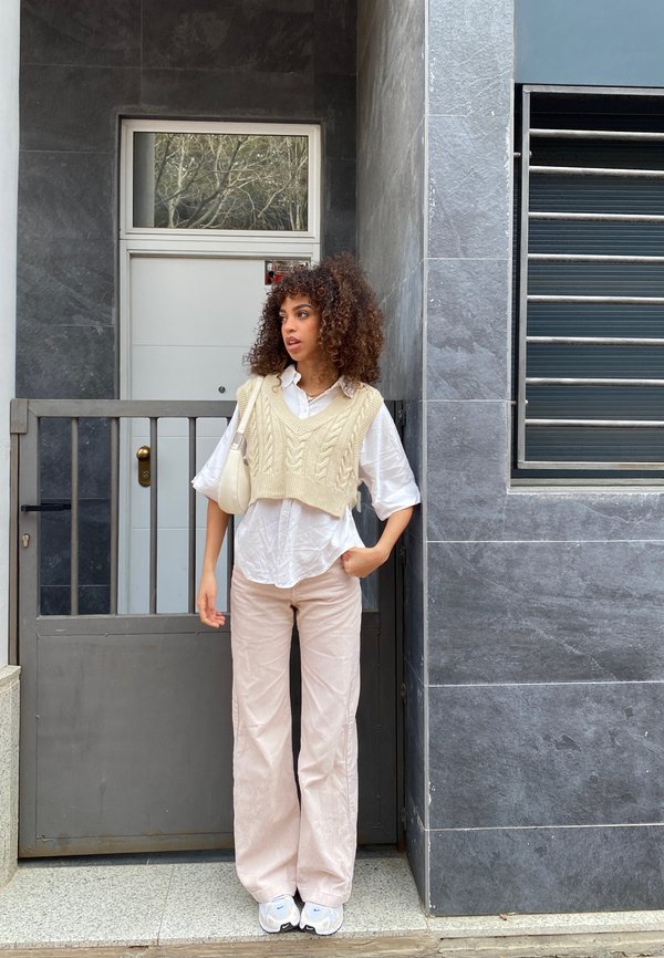 Gilet in maglia crema sopra una camicia bianca, abbinato a pantaloni in corduroy rosa chiaro e sneakers bianche. Ambientato contro un muro di pietra e una porta.