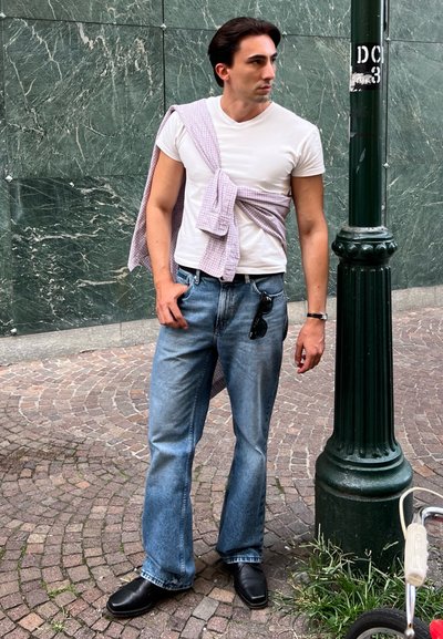 Camiseta blanca, jeans de mezclilla azul claros y zapatos negros. Camisa de cuadros morada atada a la cintura. De pie junto a un poste verde en un área de adoquines.