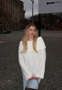 Weiße Strickjacke mit schulterfreier Gestaltung, hellblaue Jeans und einem braunen Stirnband. Hält eine braune Tasche und steht auf einer Pflasterstraße.
