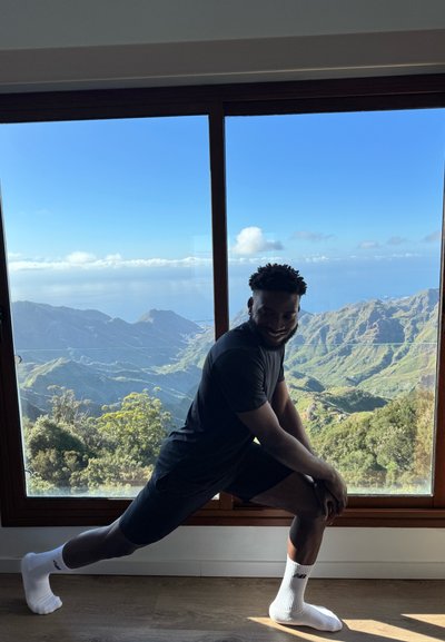 Atleta estirándose con camiseta y pantalones cortos negros, calcetines blancos, con una vista escénica de la montaña a través de una gran ventana de madera.