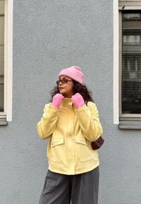 Giacca di lana gialla con due tasche frontali, abbinata a berretto rosa a coste e guanti senza dita; pantaloni grigi larghi; sfondo neutro.