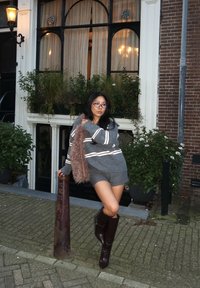 Grijze oversized trui met witte strepen, gecombineerd met grijze korte broek, bruine kniehoge laarzen en een pluizige bruine accessoire. Stedelijke omgeving met planten.