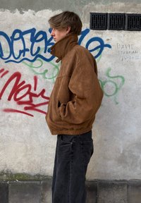 Hnědá semišová bomber bunda s vysokým límcem a elastickým pasem, doplněná černými volnými džíny, na pozadí stěny pokryté graffiti.