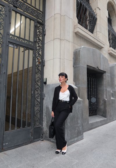 Pantalones negros y un cárdigan negro sobre una blusa blanca con encaje. Bolso negro, zapatos bicolor, y de pie contra una pared de piedra.