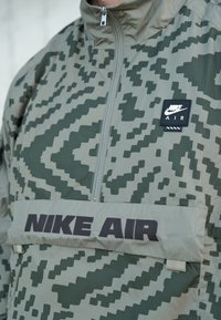 Veste coupe-vent à motif vert avec col montant, fermeture éclair avant, et bande brodée "NIKE AIR". Comprend un patch logo sur la poitrine.