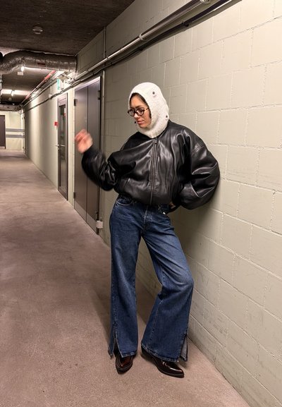 Persona con gafas que lleva una chaqueta de cuero negra, capucha de punto blanca, vaqueros azules y zapatos negros, apoyada contra una pared de ladrillos beige en un pasillo.