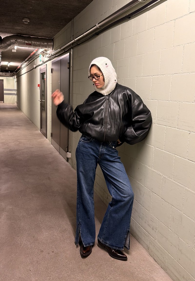 Persona con gafas que lleva una chaqueta de cuero negra, capucha de punto blanca, vaqueros azules y zapatos negros, apoyada contra una pared de ladrillos beige en un pasillo.