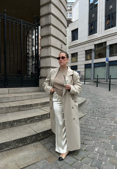 Mujer con gabardina beige, pantalones blancos de satén y gafas de sol sostiene una bebida, parada en una calle empedrada cerca de unos escalones y un edificio de piedra.